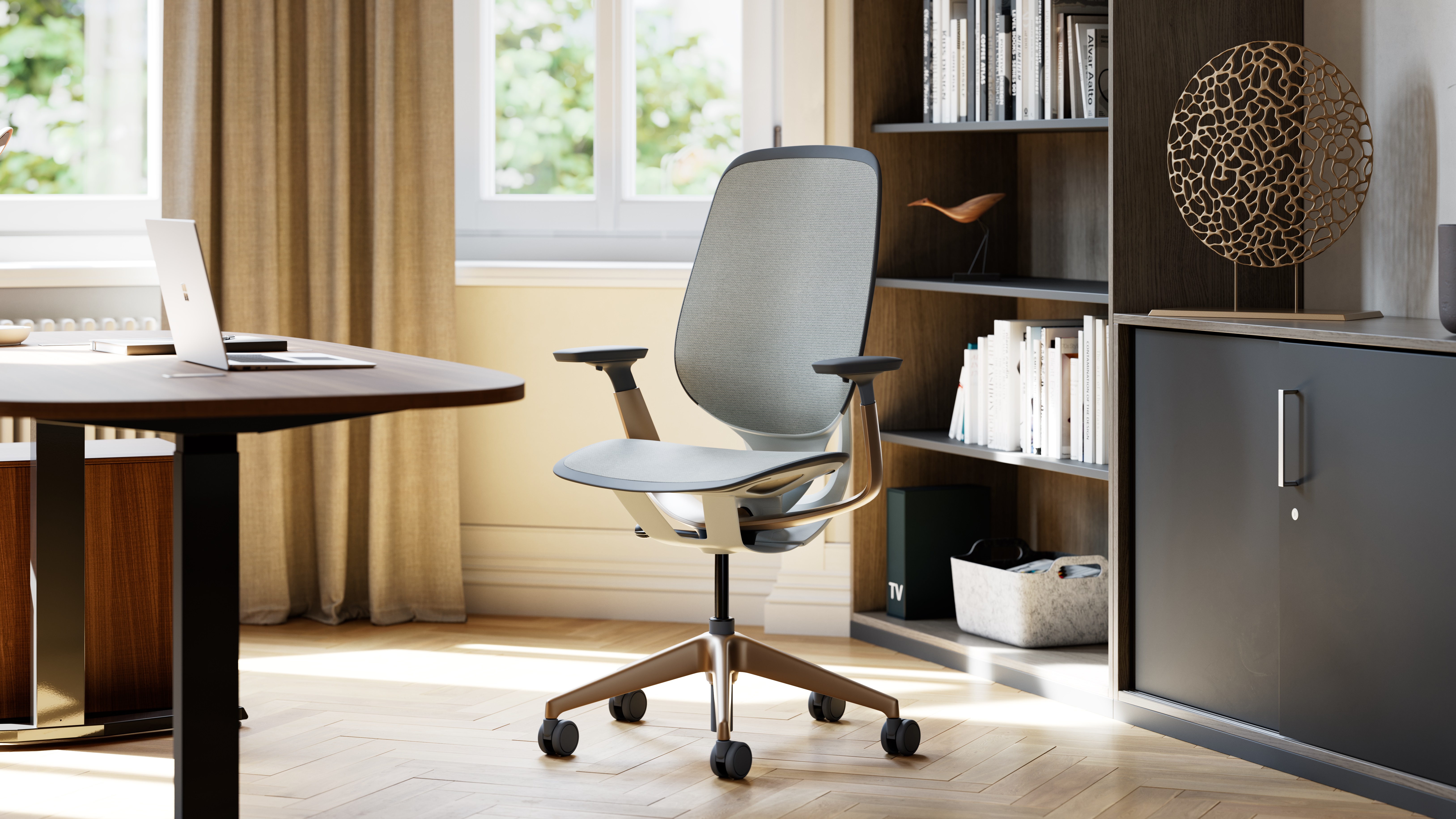 Šedá kancelářská židle Steelcase s bronzovým křížem u tmavého stolu v pracovně s knihovnou